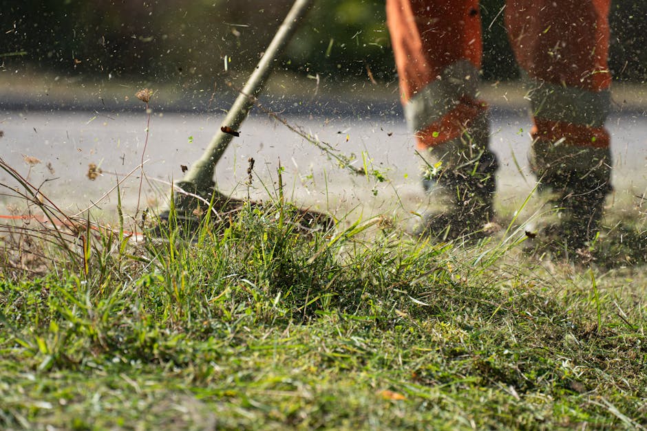 A person wearing a blue raincoat with the hood up and dark trousers is engaged in garden maintenance, using a lightweight leaf rake to clear fallen leaves and debris from a well-maintained grassy lawn